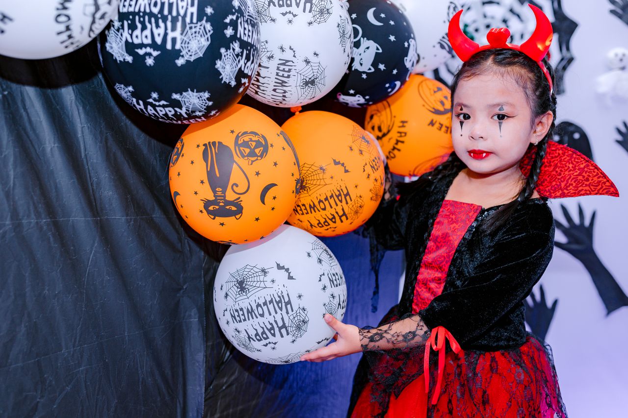 Child in devil costume holding Halloween balloons at festive celebration.